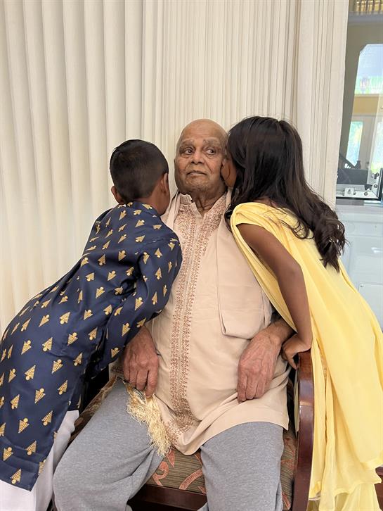 Two children lovingly kiss their grandfather while dressed in traditional festive clothing indoors.