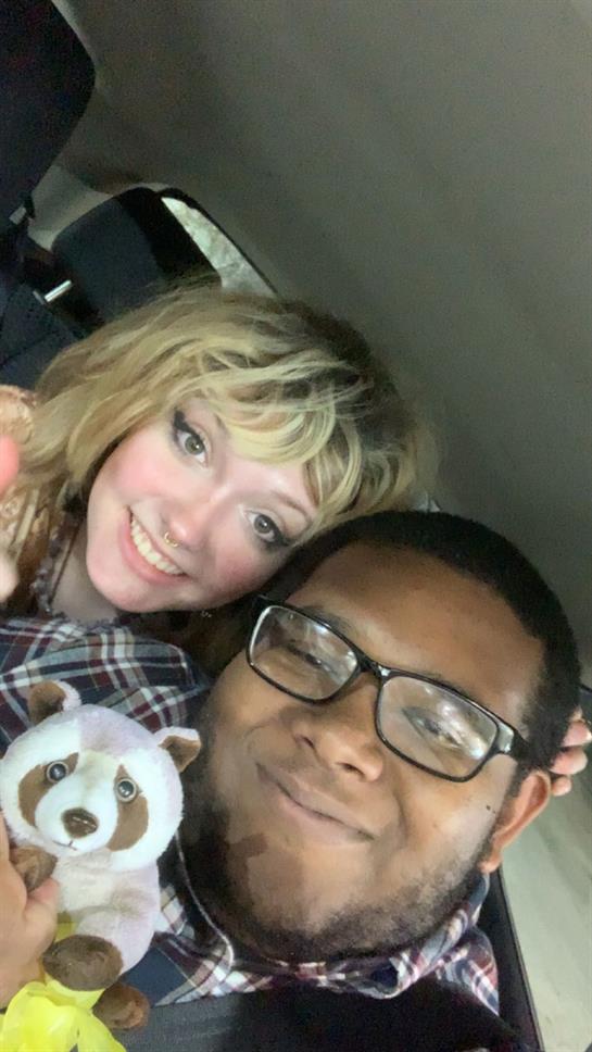 Two friends smile and pose together in a car, holding a stuffed toy and sharing a fun moment.