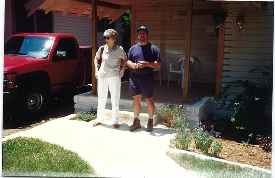 A man and woman pose on a porch in front of a house, enjoying a warm summer day.