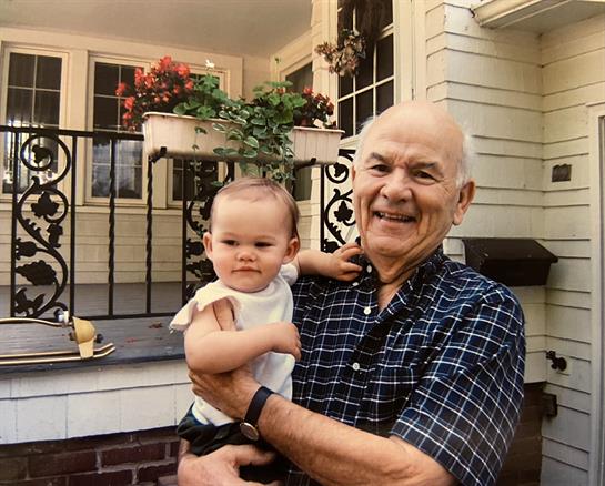 An elderly man joyfully holds a baby in a loving embrace near a well-kept house with flowers.