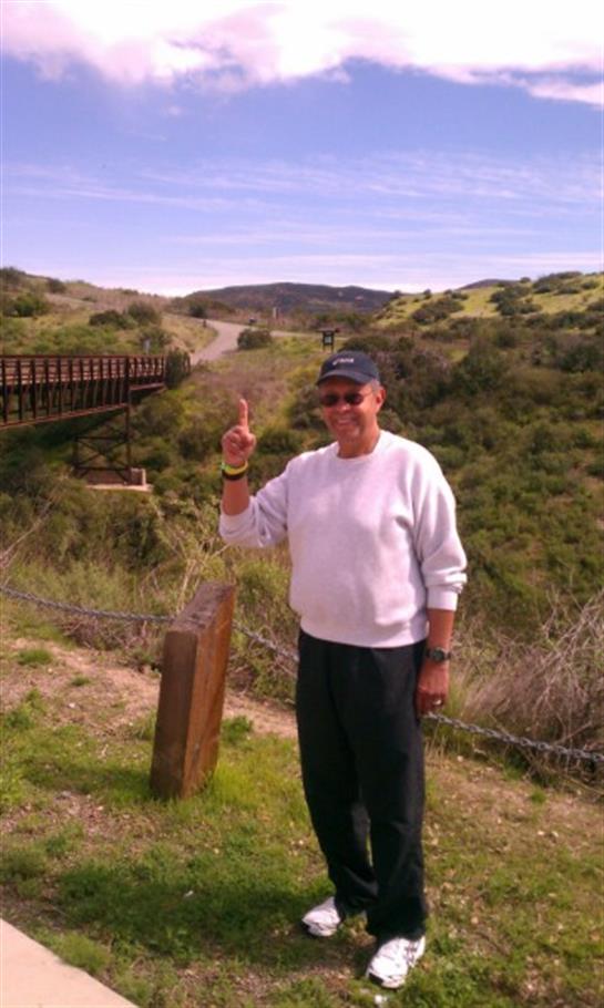 A man stands happily on a hiking trail, gesturing with one hand while surrounded by green hills.