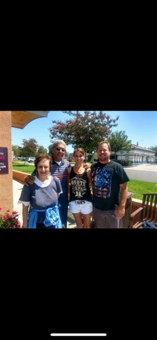 Group of four people smiling and posing together outdoors under bright sunlight in a lively area.