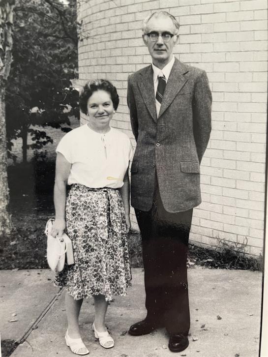 A man and woman posing together with smiles outside a light-colored building.