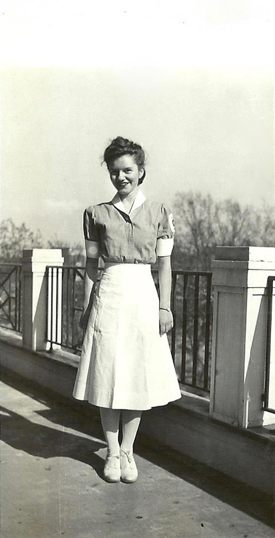 A young woman stands confidently on a balcony, wearing a vintage dress and smiling in the sunlight.