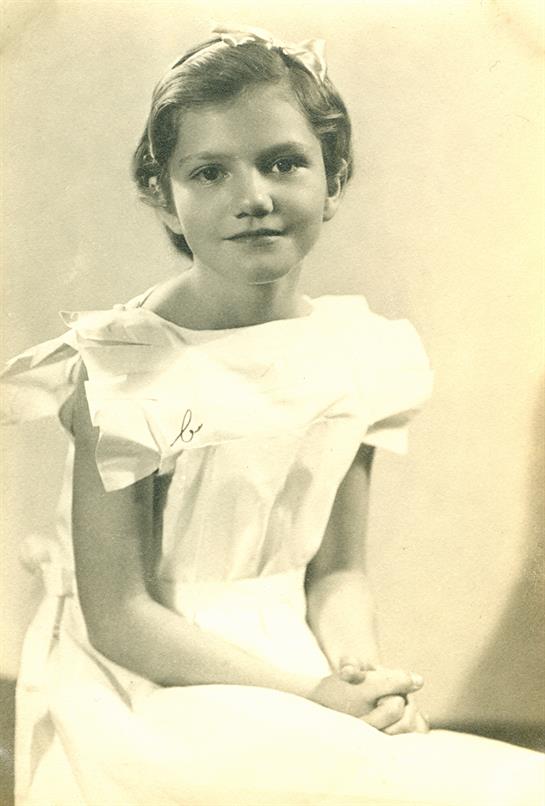 A young girl sits gracefully, wearing a vintage dress, displaying a calm demeanor in warm light.