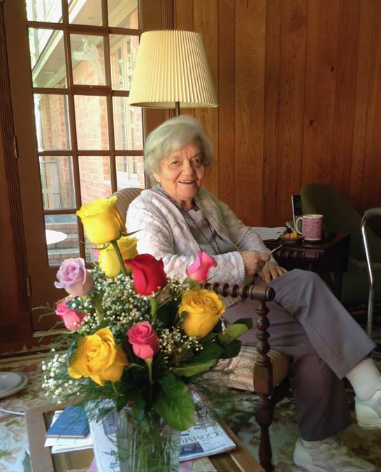A cheerful older woman relaxes in a cozy room, surrounded by a vibrant bouquet of flowers.