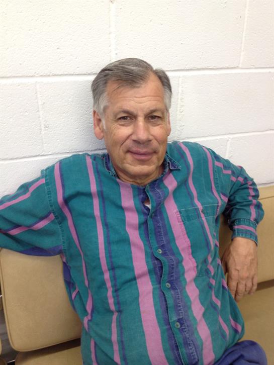 A man sits comfortably against a wall, wearing a vibrant striped shirt and smiling.