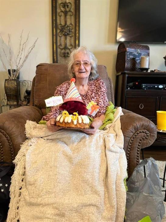 An elderly woman joyfully holds a decorated cake while celebrating her birthday in a chair.
