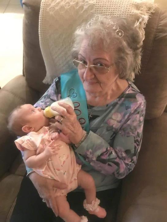 An elderly woman tenderly feeds a baby in a cozy armchair, highlighting their bond.