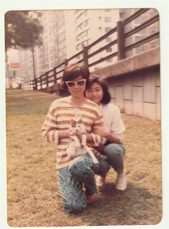 Two friends happily pose in a park with a small dog amid buildings and greenery.