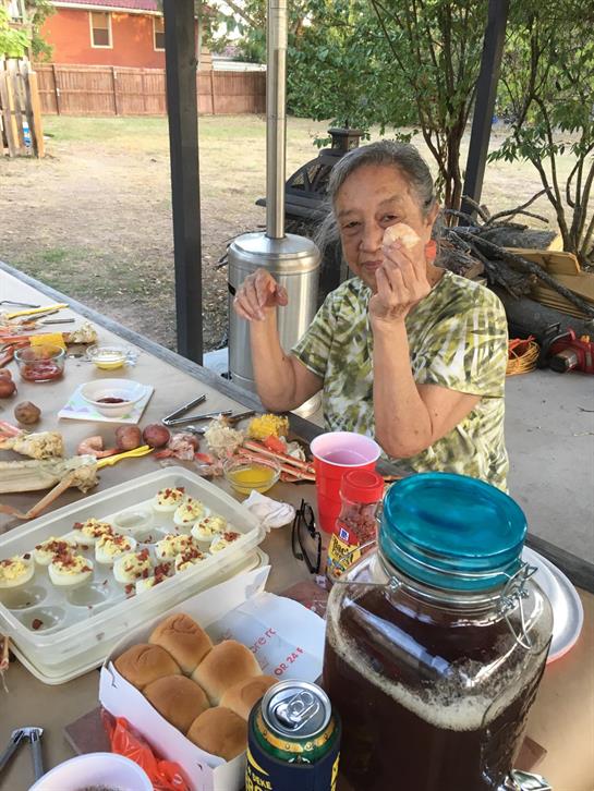 A grandmother smiles, gesturing by a table of delicious homemade food at a family gathering.