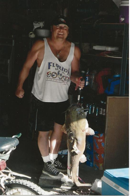 A man displays a large fish he caught during a fishing outing, smiling proudly in a garage.