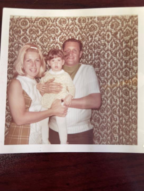 A family poses together with a child in a nostalgic home setting featuring patterned wallpaper.