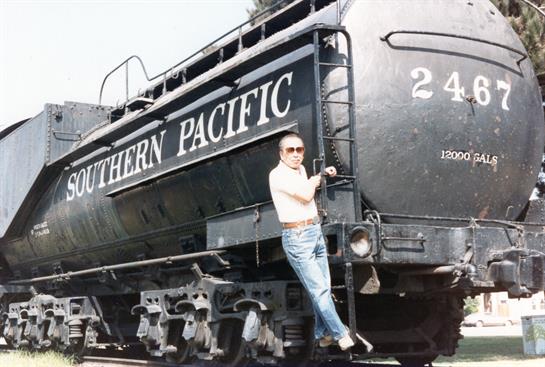 A man stands confidently on the side of an old locomotive at a railway yard under clear skies.