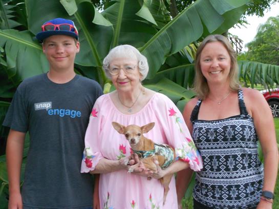 A smiling family stands together outdoors, surrounded by large green plants, with a small dog.