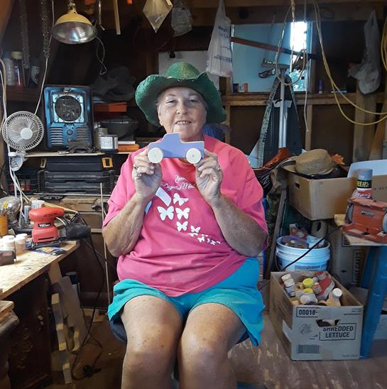 An elderly woman smiles while showing a toy car in her cluttered workshop, illuminated by sunlight.
