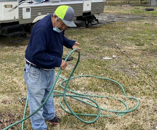 A senior man is coiling a green garden hose in the grass by a camper, preparing for work.