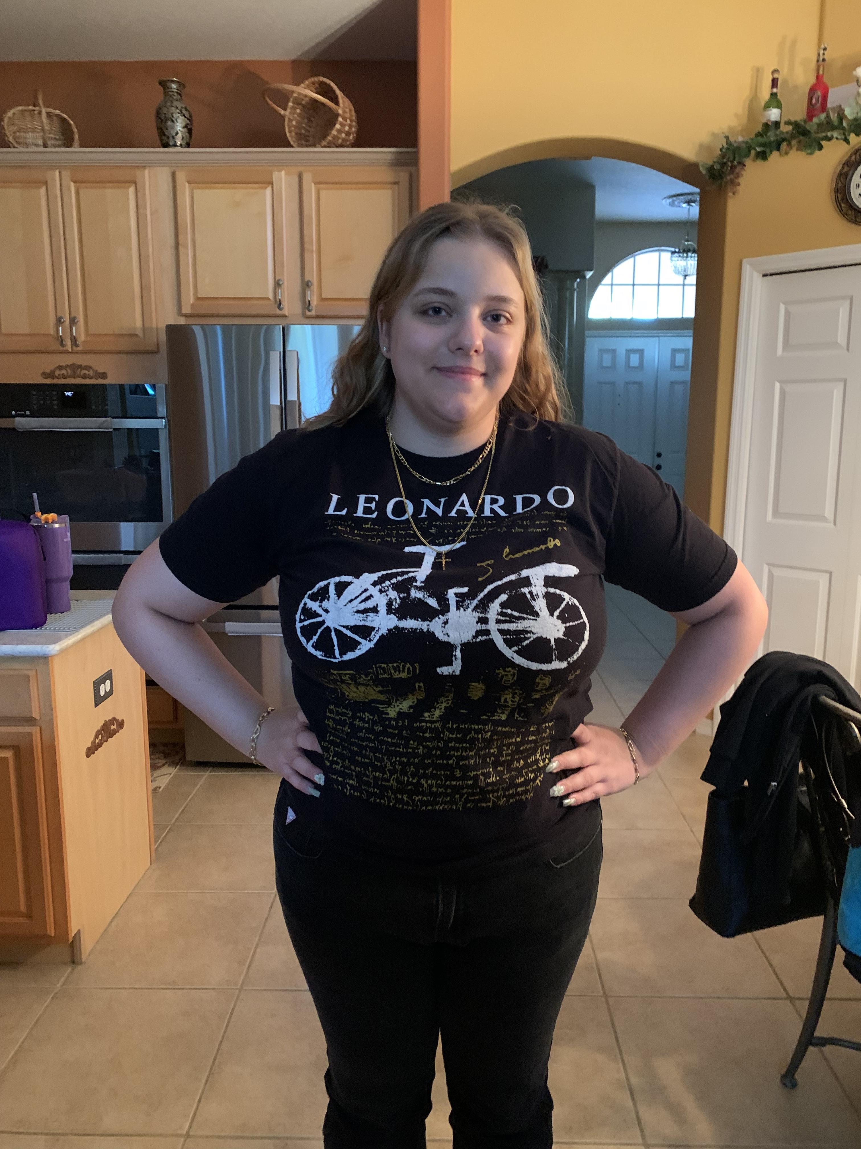 A young woman smiles with hands on hips in a bright kitchen with wooden cabinets.