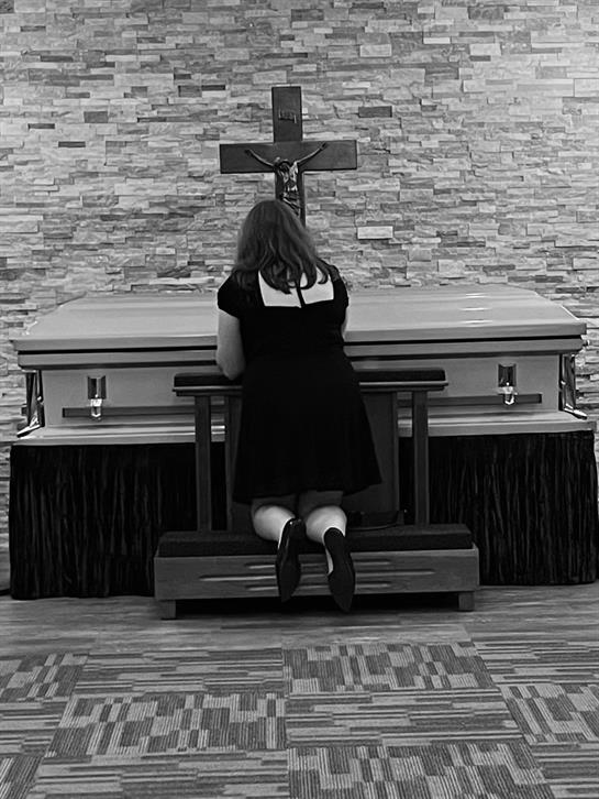 A woman is kneeling in front of a memorial altar, reflecting in a solemn atmosphere.