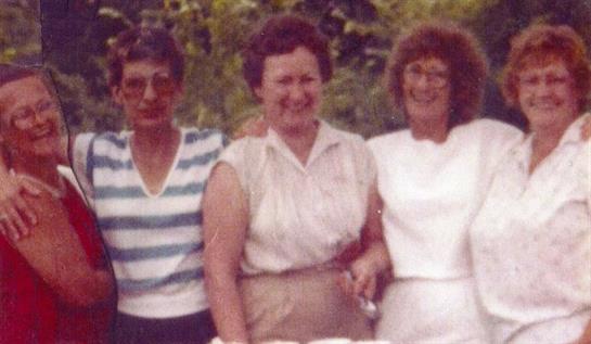 Three women smile while standing outdoors, enjoying a sunny afternoon in a garden.