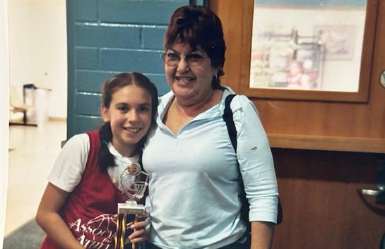 A young girl proudly holds a trophy beside a smiling woman. They are at a community event.