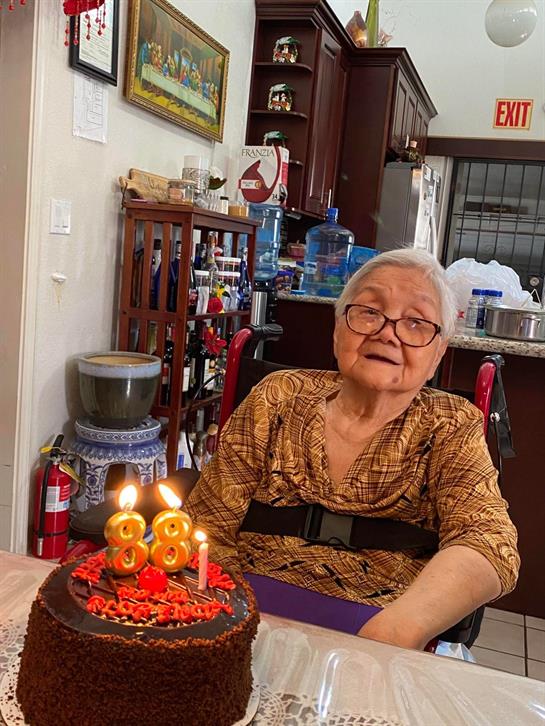Elderly woman sits at a table, joyfully celebrating her birthday with cake featuring lit candles.