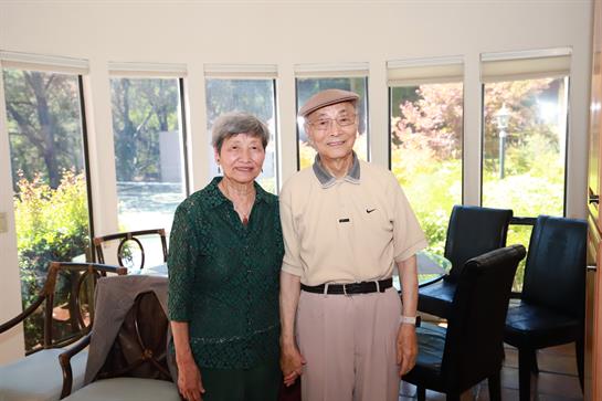 An elderly couple stands hand in hand, smiling warmly at each other, surrounded by light.