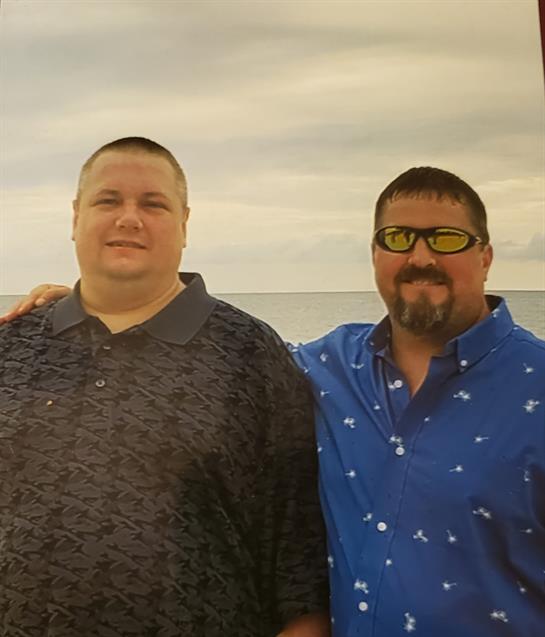 Two men stand together on a beach, smiling as they enjoy a sunny day by the water.