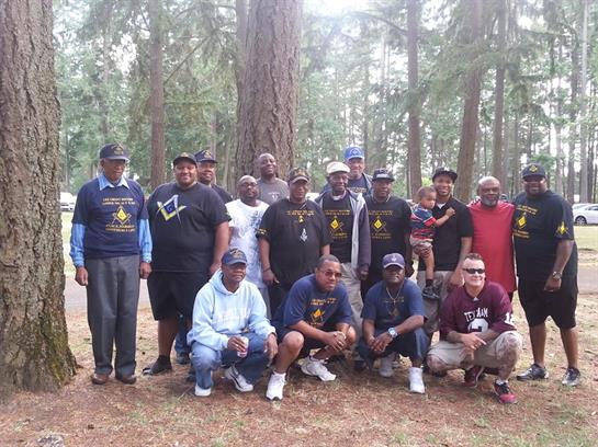Men of various ages stand and sit together in a forest clearing, enjoying a reunion during daylight.