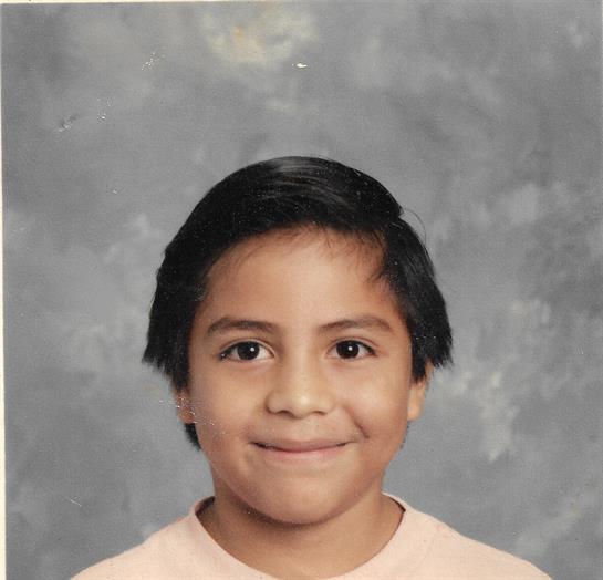 A young boy smiles in a school portrait, showing short black hair and wearing a light-colored shirt.