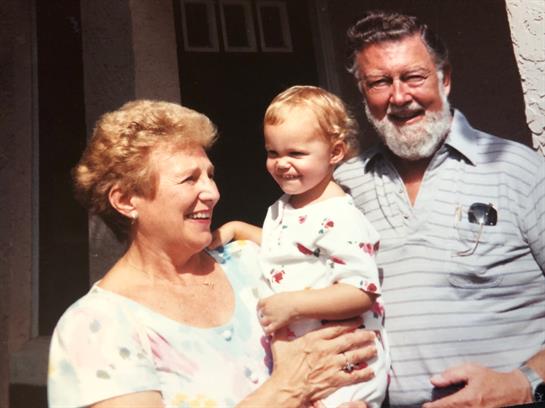 Grandparents smiling while holding their laughing grandchild in a cheerful outdoor setting.