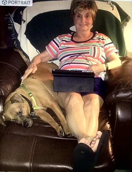 A woman sits comfortably on a couch with a dog resting beside her as she uses a tablet.