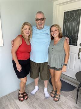 Three friends smile outside a house on a warm, sunny day, enjoying their time together.