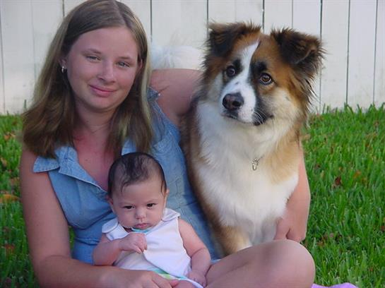 Mother holds her baby while sitting on grass alongside their friendly dog, all enjoying a warm day.