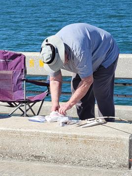 An older man organizes his fishing gear by the water, enjoying a calm day outdoors.