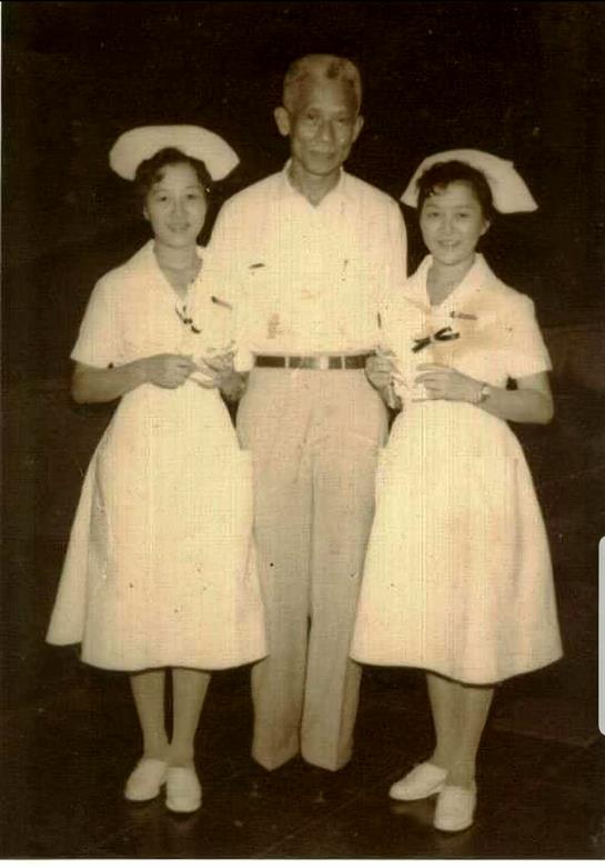 Two nurses in vintage uniforms stand beside a man, all smiling and holding flowers.