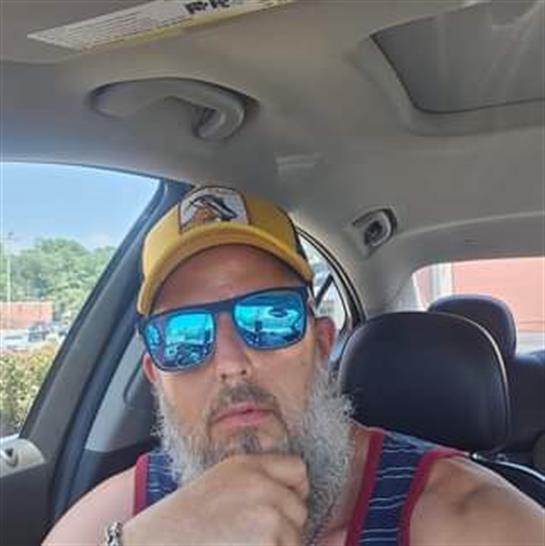 A man with a beard is seated in a car, wearing sunglasses and a cap while thinking.