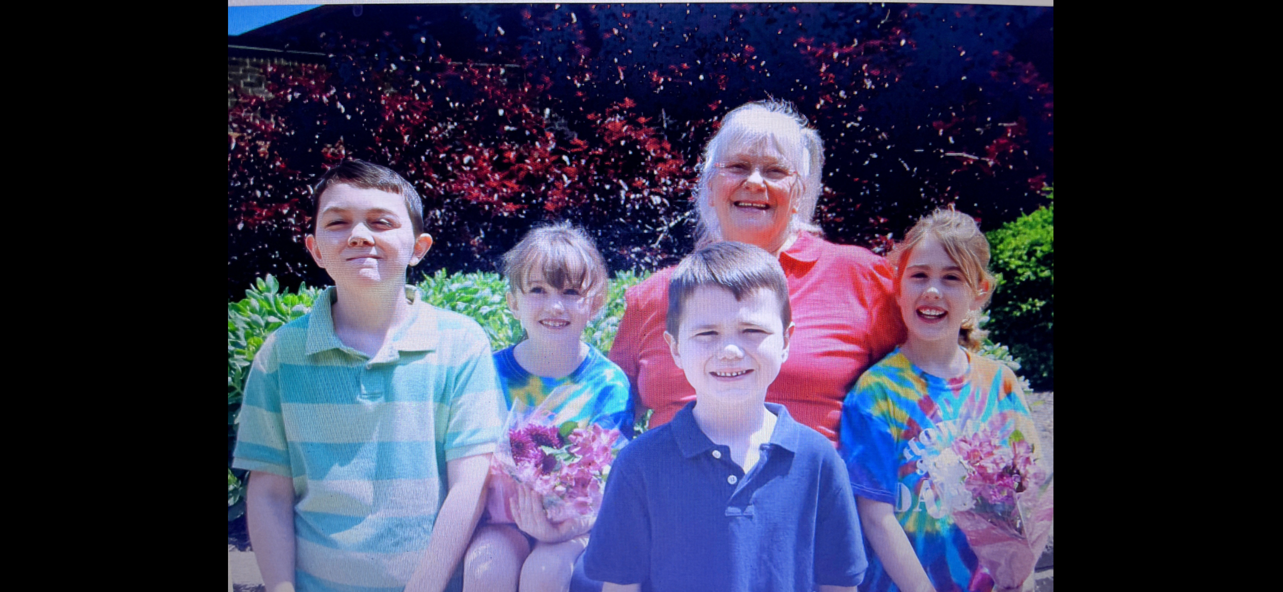 Children smile brightly while posing with their grandmother in a vibrant outdoor garden setting.
