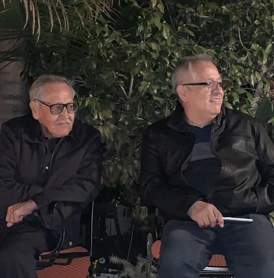 Two men in black jackets casually converse while seated outdoors under greenery during the evening.