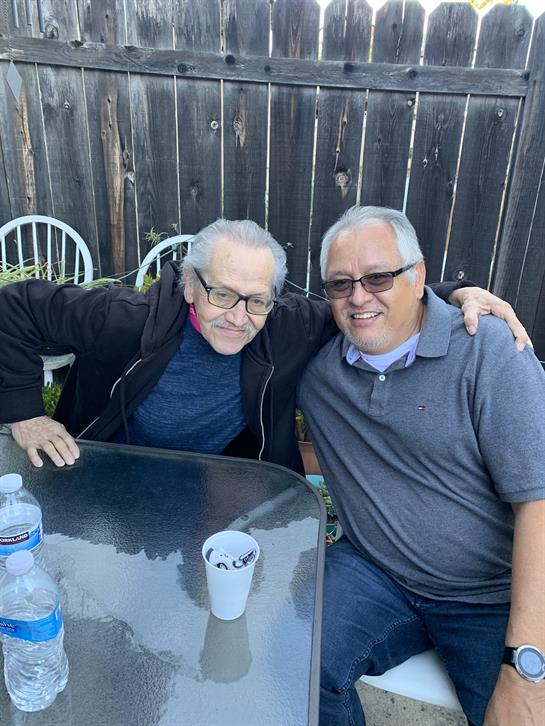 Two men sit close at a table, smiling and enjoying a casual conversation in a cozy backyard.