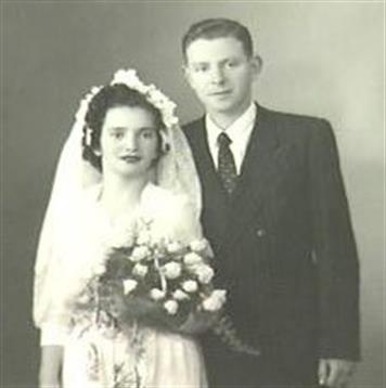 Newlywed couple stands together, dressed in wedding attire with flowers, in a classic vintage style.