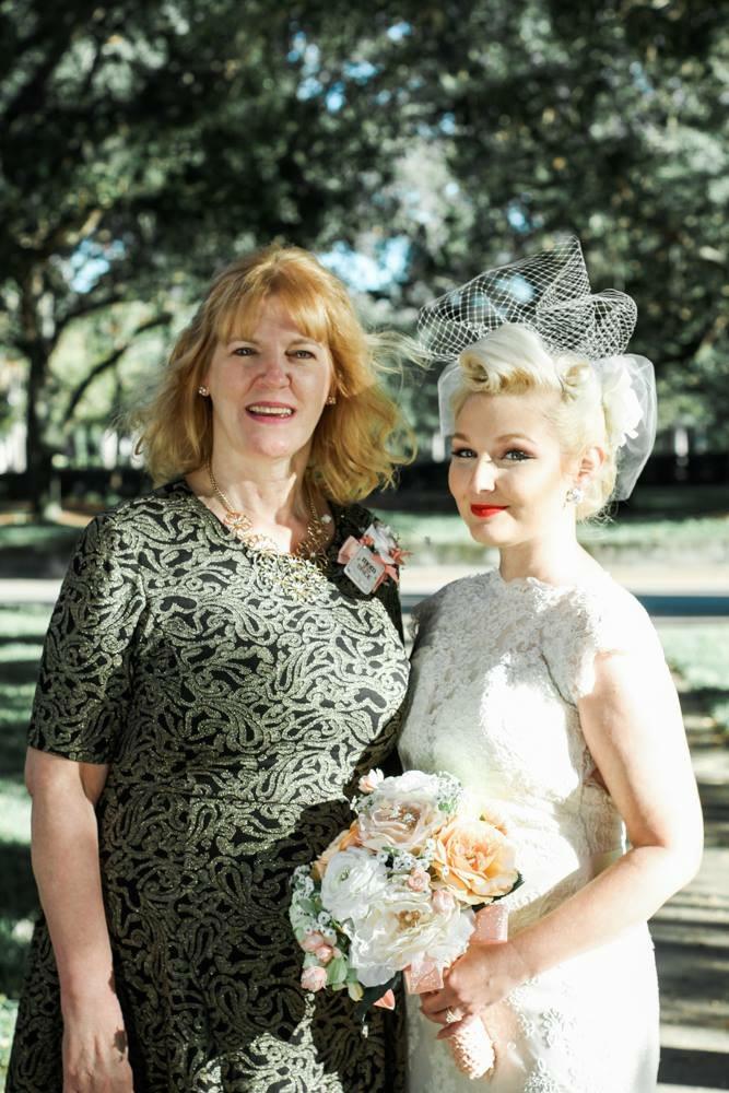 Two women outdoors, in elegant dresses, holding a bouquet and smiling happily.