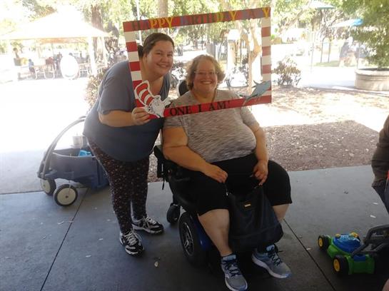 Two women pose joyfully inside a festive frame at a park event, enjoying the sunny day.