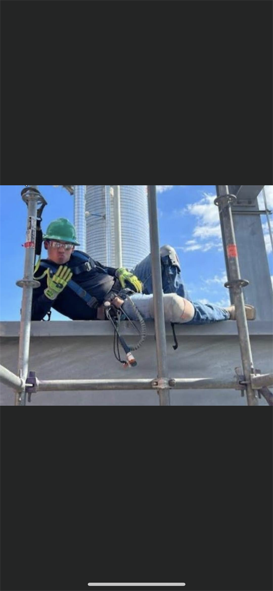 A construction worker relaxes on a scaffold, displaying safety gear against a bright sky.