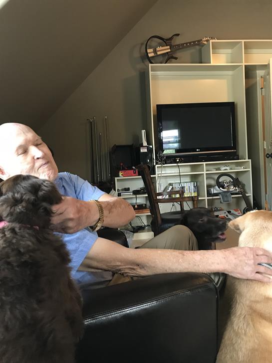 An older man sits in a cozy home, affectionately petting his two dogs.