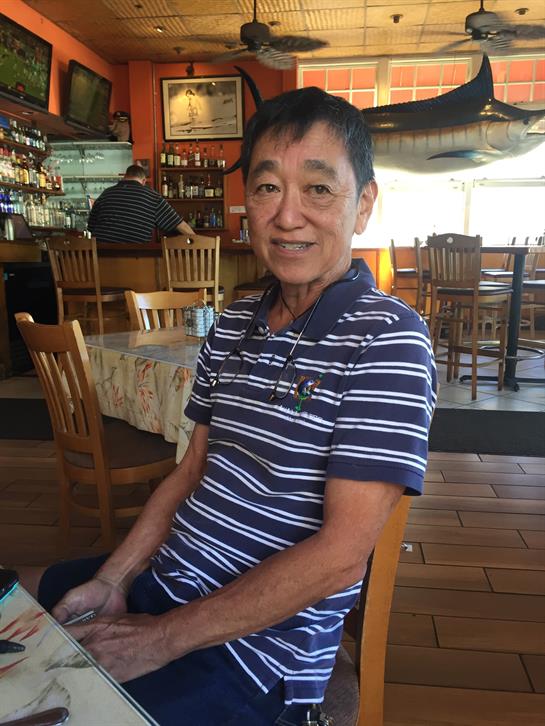 An older man in a striped shirt relaxes at a cafe table, smiling contently while dining.