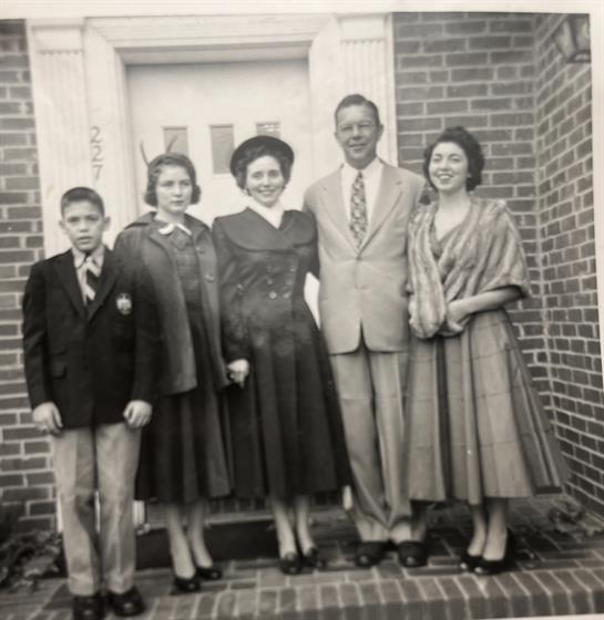 Five family members pose together outside a brick house, dressed in formal attire for a celebration.