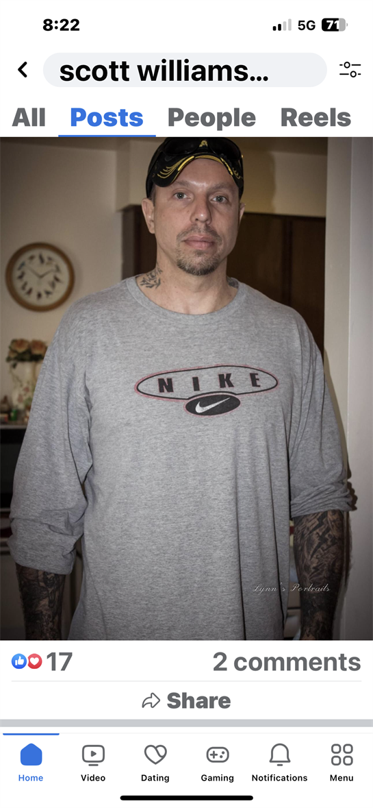 Male individual poses indoors in a gray shirt, showcasing tattoos on his arms.