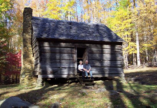 Two people relax on the cabin steps surrounded by colorful autumn foliage in the forest.