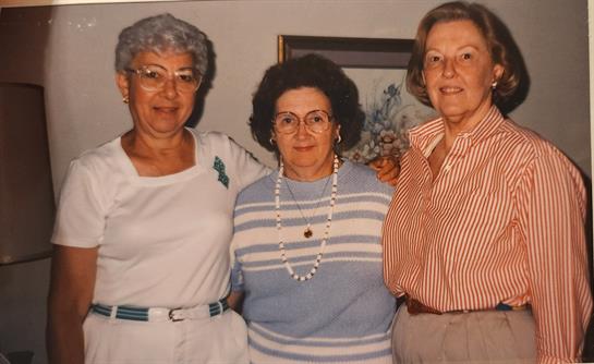 Three women stand together, smiling happily in a warm indoor environment during a casual gathering.